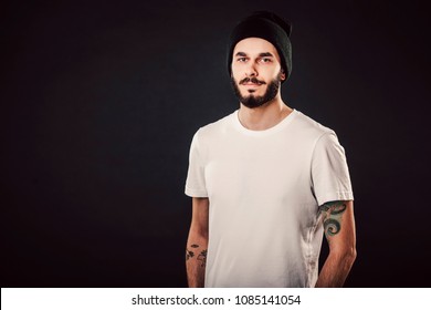 Waist up portrait of serious stylish attractive man with thick beard looks confidently, dressed in casual white t shirt, isolated over black background. Young hipster in cap with tattoo. Mock up