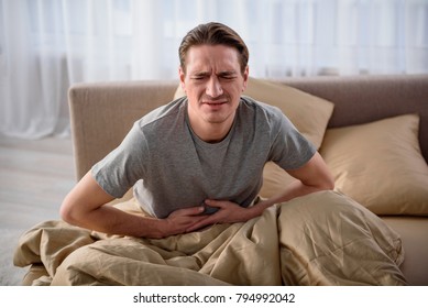 Waist up portrait of sick man sitting on bed with closed eyes. He is terribly ill and having a stomachache