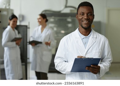 Waist up portrait of smiling black man wearing lab coat looking at camera in factory workshop and holding clipboard, copy space - Powered by Shutterstock