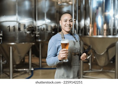 Waist up portrait of smiling female brewmaster holding beer glass and looking at camera while working at brewing factory - Powered by Shutterstock