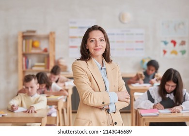 Waist up portrait of smiling female teacher looking at camera while posing confidently standing with arms crossed in school classroom, copy space - Powered by Shutterstock