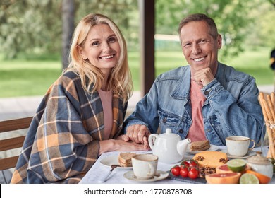 Waist up portrait of smiling loving spouses sitting at table with tea and snacks on terrace in countryside - Powered by Shutterstock