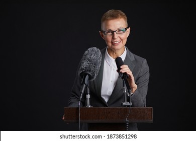 Waist Up Portrait Of Smiling Mature Woman Standing At Podium And Speaking To Microphone Against Black Background, Copy Space