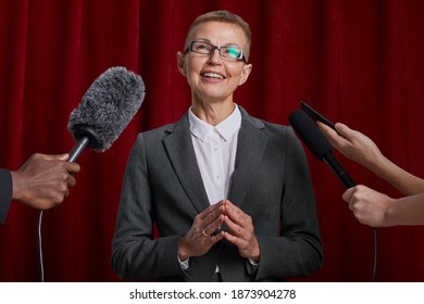 Waist Up Portrait Of Smiling Mature Woman Giving Interview To Journalist And Speaking To Microphones Against Red Curtain