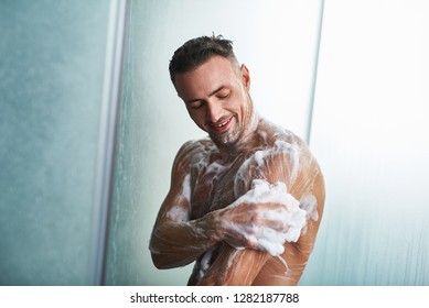 Waist up portrait of smiling naked gentleman with foam on his body taking shower at home