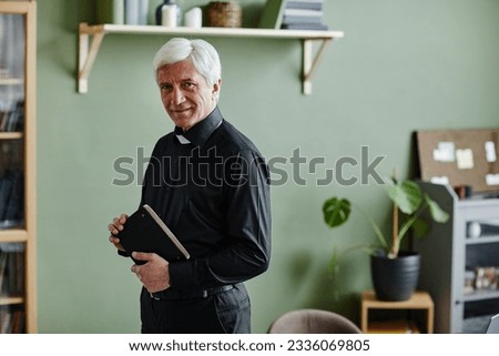 Waist up portrait of smiling senior priest looking at camera and holding Bible while standing in office against green wall, copy space