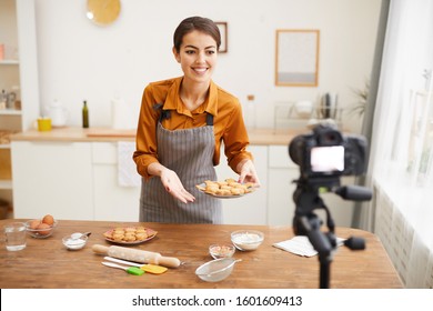 Waist up portrait of smiling young woman holding homemade cookies while filming baking tutorial for video channel, copy space