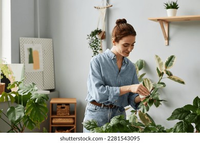 Waist up portrait of smiling young woman caring for lush greenery at home and inspecting plants - Powered by Shutterstock