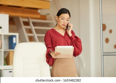 Waist up portrait of successful Asian businesswoman speaking by phone and holding clipboard while working in modern white office, copy space