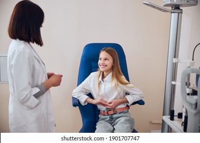 Waist Up Portrait Of Teenager Girl Sitting In Blue Medicine Chair While Having Conversation With Experienced Female Ophthalmologist In Front Of Her In Optician Cabinet