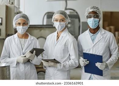 Waist up portrait of three factory workers wearing lab coats and masks looking at camera in workshop - Powered by Shutterstock