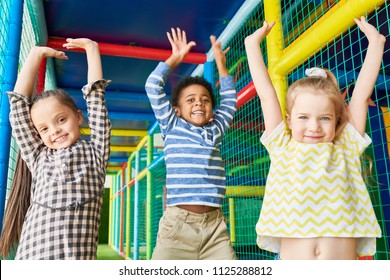 Waist up portrait of three happy kids jumping raising hands while having fun in play area and looking at camera