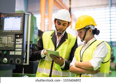 Waist up portrait of two Asian factory workers with safety helmet check stock in digital tablet near monitor of industrial machine. Manufacturing industry to produce car parts. Quality assurance (QA).
