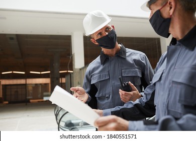 Waist Up Portrait Of Two Construction Workers Wearing Masks And Discussing Plans While Standing In Shopping Mall Or Office Building, Copy Space