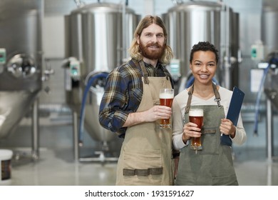 Waist up portrait of two smiling young workers holding beer glasses and looking at camera while standing in workshop at brewing factory, copy space - Powered by Shutterstock