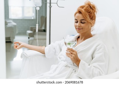 Waist up portrait view of the charming woman in white bathrobe sitting in armchair and receiving IV infusion. She is holding glass of lemon beverage and smiling 