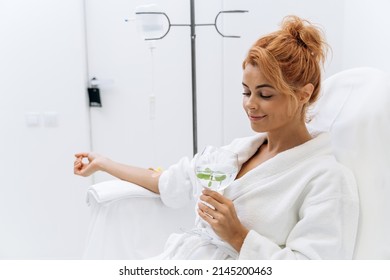 Waist up portrait view of the charming woman in white bathrobe sitting in armchair and receiving IV infusion. She is holding glass of lemon beverage and smiling 