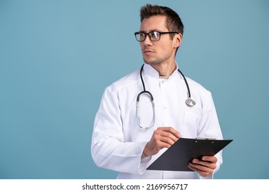 Waist Up Portrait View Of The Confident Doctor In A Uniform And In Glasses Holding Folder And Looking Away With Serious Face. Stock Photo 