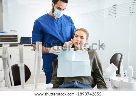 Waist up portrait view of the multiracial serious doctor carefully putting a protective towel on his female patient sitting at the dentist armchair. People, medicine, stomatology and health care