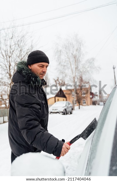 Waist up portrait view of the\
senior man cleaning snow from car windshield outdoors on winter\
day. Male person with cigarette cleaning window of his\
car