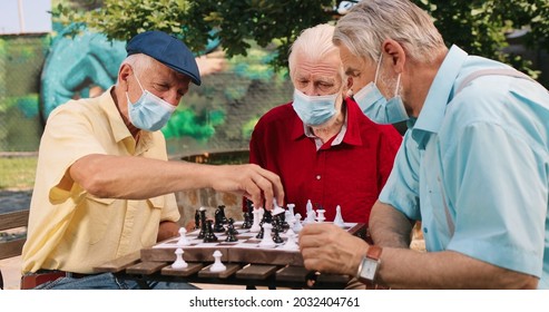 Waist Up Portrait View Of The Three Caucasian Old Retired And Smiled Men Wearing Protective Masks Emotionally Chatting And Playing At The Chess. Outside. Leisure Games Concept 