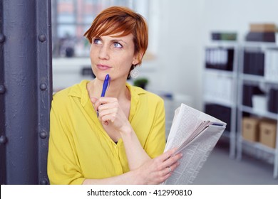 Waist Up Portrait of Woman with Short Red Hair Leaning Against Pole in Modern Home Office Holding Pen and Newspaper and Looking Deep in Thought - Doing Crossword or Reading Ads