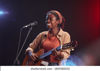 Waist up portrait of young African-American woman playing guitar on stage and singing to microphone smiling, copy space - Powered by Shutterstock