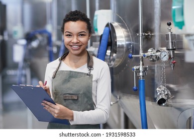 Waist up portrait of young African-American woman enjoying work at industrial factory and smiling at camera holding clipboard, copy space - Powered by Shutterstock