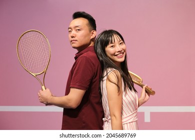 Waist up portrait of young asian couple holding tennis racket while posing back to back against pastel pink background. - Powered by Shutterstock