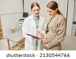 Waist up portrait of young female doctor consulting pregnant woman in clinic and using digital tablet