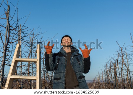 Waist up portrait of the young happy man gardener, dressed in black hat, blue jacket and orange work gloves stands with closed eyes among his garden near the wooden stepladder and shows rock and roll
