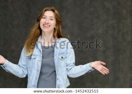 Waist up portrait of a young pretty brunette girl woman with beautiful long hair on a gray background in a jeans jacket. He talks, smiles, shows his hands with emotions in various poses.