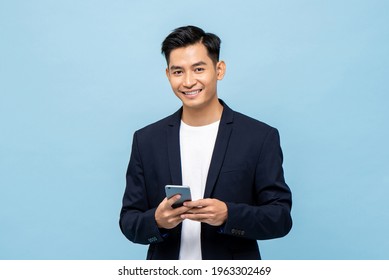 Waist up portrait of Young smiling handsome Asian man in semi formal suit using mobile phone in light blue isolated studio background - Shutterstock ID 1963302469