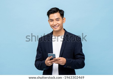 Waist up portrait of Young smiling handsome Asian man in semi formal suit using mobile phone in light blue isolated studio background
