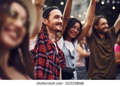 Waist Up Portrait Of Young Stylish Man Raising Hands And Looking Away With Smile. Group Of Hipsters On Blurred Background