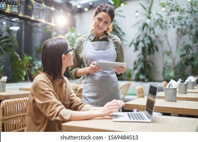 Waist up portrait of young waitress holding digital tablet while taking order of clients at outdoor cafe terrace decorated with plants, copy space