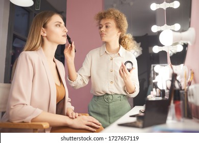 Waist Up Portrait Of Young Woman Applying Make Up On Fashion Model By Vanity Mirror In Pink Dressing Room Interior, Copy Space