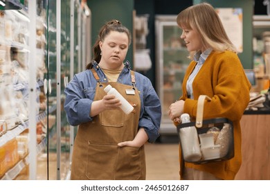 Waist up portrait of young woman with Down syndrome working as assistant in supermarket and consulting customer in dairy aisle - Powered by Shutterstock