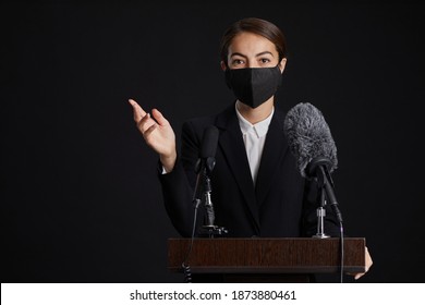 Waist Up Portrait Of Young Woman Wearing Mask While Giving Speech Standing At Podium Against Black Background, Copy Space