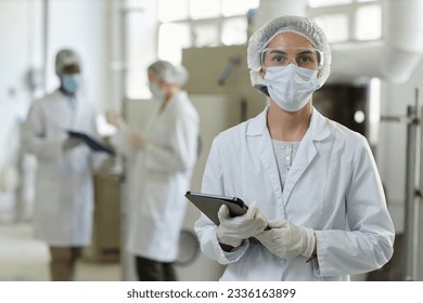 Waist up portrait of young woman wearing mask and lab coat looking at camera in factory workshop, copy space - Powered by Shutterstock