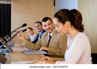 Waist up profile view of panel of business colleagues discussing amongst each other in conference room in front of visual screens and microphones.