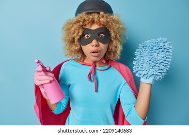 Waist Up Shot Of Amazed Scared Curly Female Model Dressed In Superhero Costume Holds Detergent And Rag On Hand Washes Something Dirty At Home Isolated Over Blue Background. Housework Concept