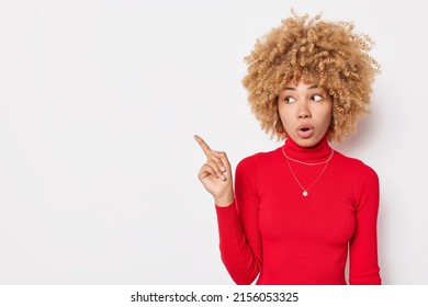 Waist up shot of amazed woman with curly hair points away at blank space feels shocked wears red turtleneck isolated over white background. Curious female asks question about product poses indoor