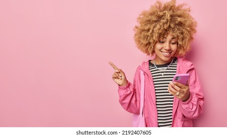 Waist Up Shot Of Beautiful Curly Haired European Woman Dressed Casually Holds Mobile Phone Checks Received Message Points At Blank Copy Space Isolated Over Pink Background Advertises Product