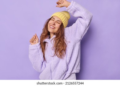 Waist up shot of carefree young European woman with long hair daces and catches every bit of music smiles broadly keeps eyes closed dressed in winter outerwear isolated over purple background