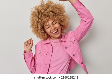 Waist Up Shot Of Cheerful European Woman With Blonde Curly Hair Dances Carefree Shakes Arms Has Upbeat Mood Dressed In Pink Jacket Smiles Broadly Isolated Over Grey Background. Winner Dance.