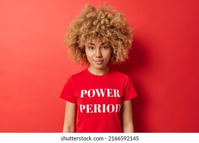 Waist Up Shot Of Curly Haired Young European Woman Looks Directly At Camera Has Calm Expression Dressed In Casual T Shirt With Inscription Isolated Over Vivid Red Background. Monochrome Shot