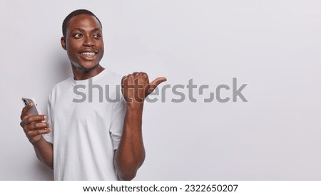 Waist up shot of dark skinned man smiles positively holds smartphone pointing aside with his thumb recommends fantastic offer inviting you to explore exciting opportunity showcased in copy space