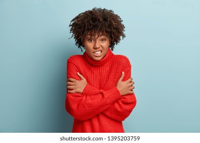 Waist up shot of dissatisfied dark skinned woman crosses hands over chest, feels scared, trembles from fear, frowns face, wears red warm sweater, isolated over blue background. Its chilling there - Shutterstock ID 1395203759