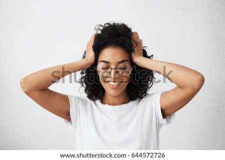 Waist up shot of excited amazed young mixed race woman in eyewear touching her black curly hair and keeping eyes closed, feeling absolutely happy after her boyfriend finally proposed to her